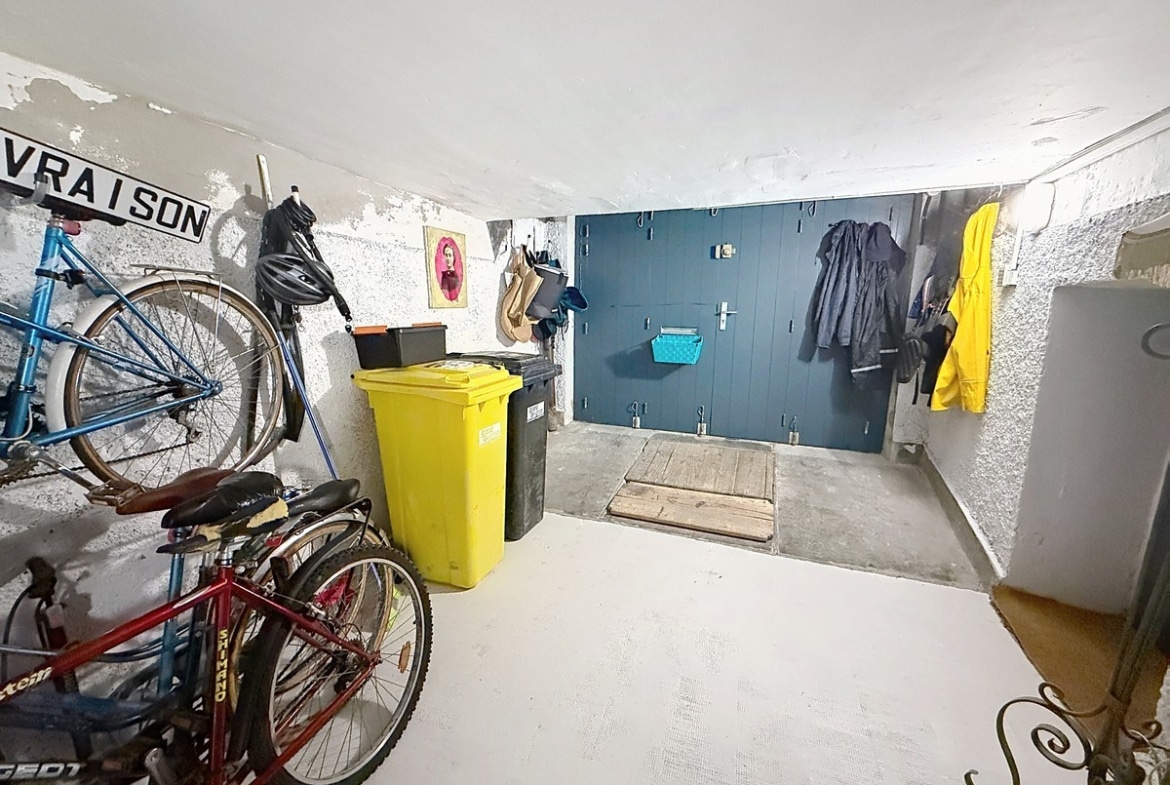 Local de rangement avec vélos suspendus au mur, poubelles jaune et noire, et placard mural bleu pétrole. Sol béton gris, murs en crépi blanc, vestes imperméables accrochées et trappe au sol.