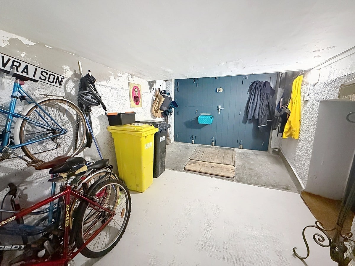 Local de rangement avec vélos suspendus au mur, poubelles jaune et noire, et placard mural bleu pétrole. Sol béton gris, murs en crépi blanc, vestes imperméables accrochées et trappe au sol.