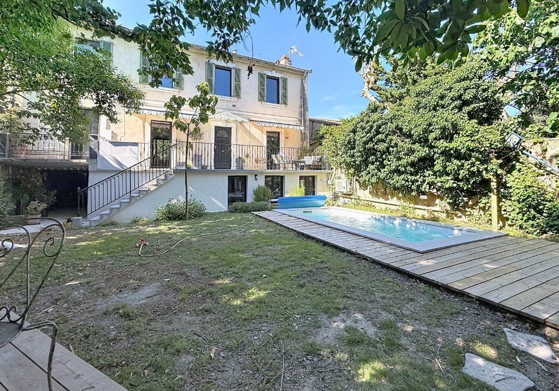 Jardin arboré avec pelouse et piscine rectangulaire entourée d'une terrasse en bois composite. Façade arrière de maison de caractère sur deux niveaux avec volets verts, escalier extérieur en fer forgé et terrasse surélevée.