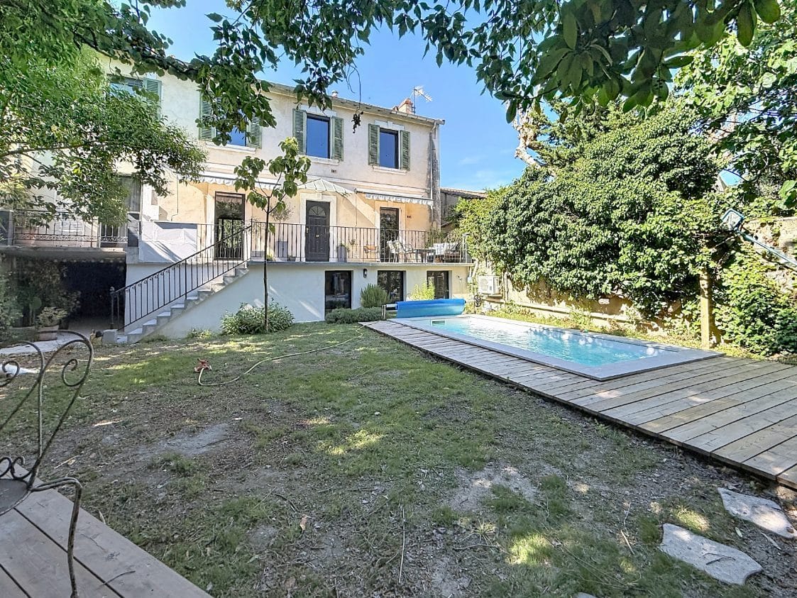 Jardin arboré avec pelouse et piscine rectangulaire entourée d'une terrasse en bois composite. Façade arrière de maison de caractère sur deux niveaux avec volets verts, escalier extérieur en fer forgé et terrasse surélevée.