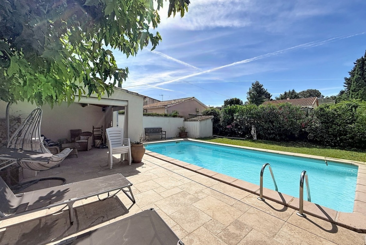 Piscine rectangulaire bleu turquoise avec échelle chromée, entourée de dalles beiges. Transats gris au premier plan, pool house couvert avec mobilier de jardin blanc et pergola en bois.