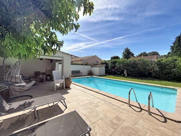 Piscine rectangulaire bleu turquoise avec échelle chromée, entourée de dalles beiges. Transats gris au premier plan, pool house couvert avec mobilier de jardin blanc et pergola en bois.