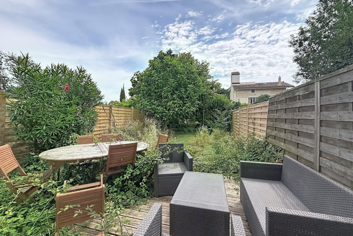 Jardin privatif clôturé par des panneaux de bois horizontaux avec terrasse en lames de bois. Mobilier de jardin comprenant une table ronde en teck avec chaises et un salon bas en résine tressée grise, entouré de végétation dense incluant lauriers roses et arbres fruitiers.