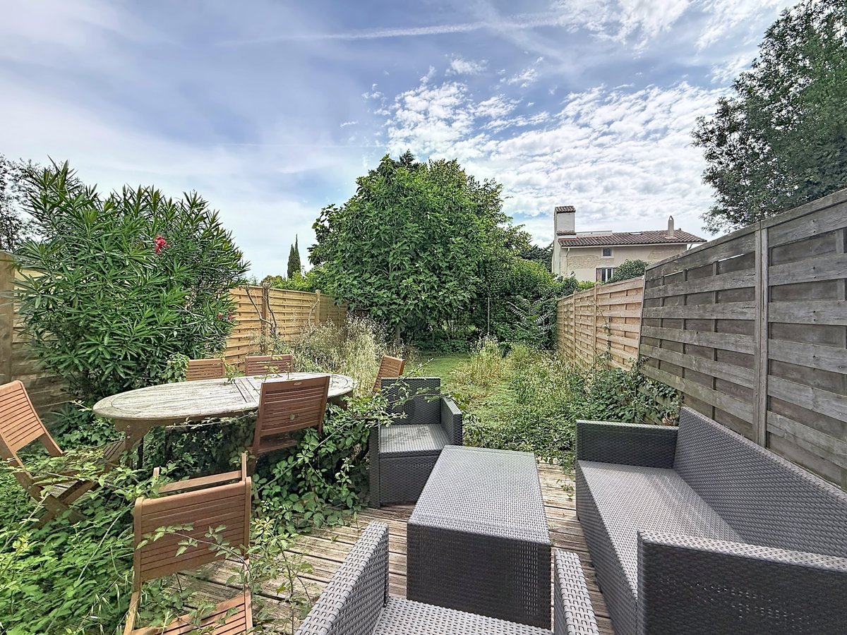 Jardin privatif clôturé par des panneaux de bois horizontaux avec terrasse en lames de bois. Mobilier de jardin comprenant une table ronde en teck avec chaises et un salon bas en résine tressée grise, entouré de végétation dense incluant lauriers roses et arbres fruitiers.