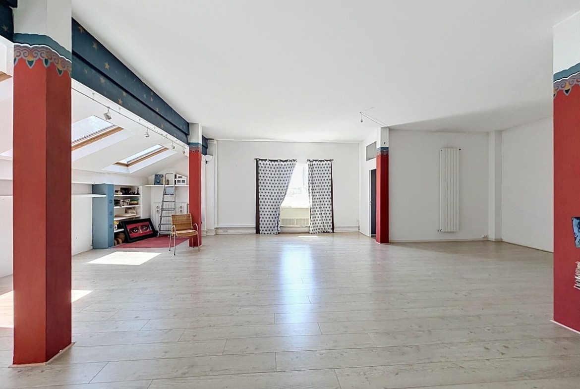 Grande pièce sous combles avec parquet clair et poutres peintes en bleu nuit étoilé. Colonnes décoratives rouges ornées de motifs, trois velux, radiateur vertical blanc, rideaux à pois.