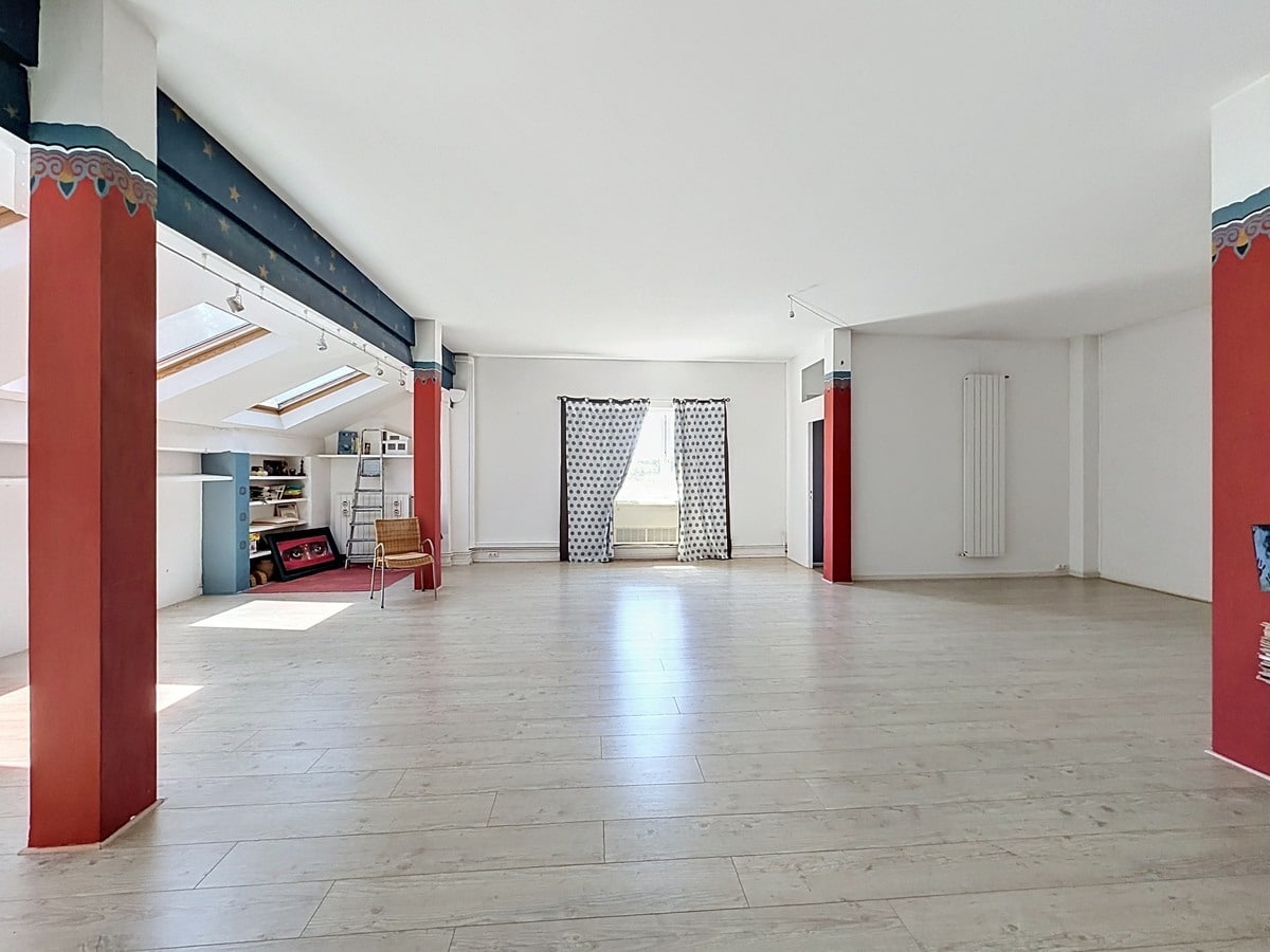 Grande pièce sous combles avec parquet clair et poutres peintes en bleu nuit étoilé. Colonnes décoratives rouges ornées de motifs, trois velux, radiateur vertical blanc, rideaux à pois.
