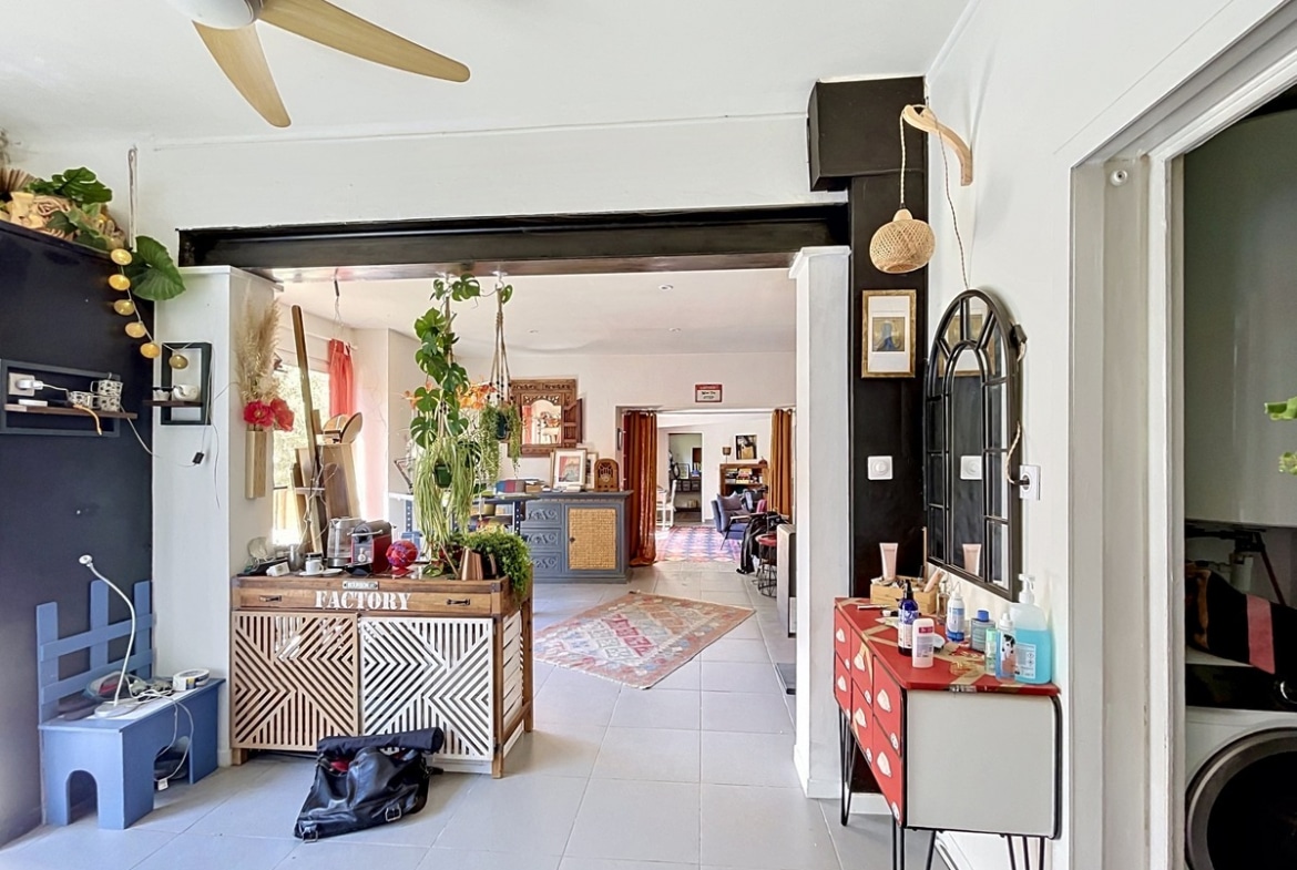 Espace de vie ouvert avec ventilateur de plafond en bois et poutre métallique noire apparente. Buffet en bois à motifs géométriques, commode rouge à tiroirs, plantes suspendues et carrelage gris clair au sol.