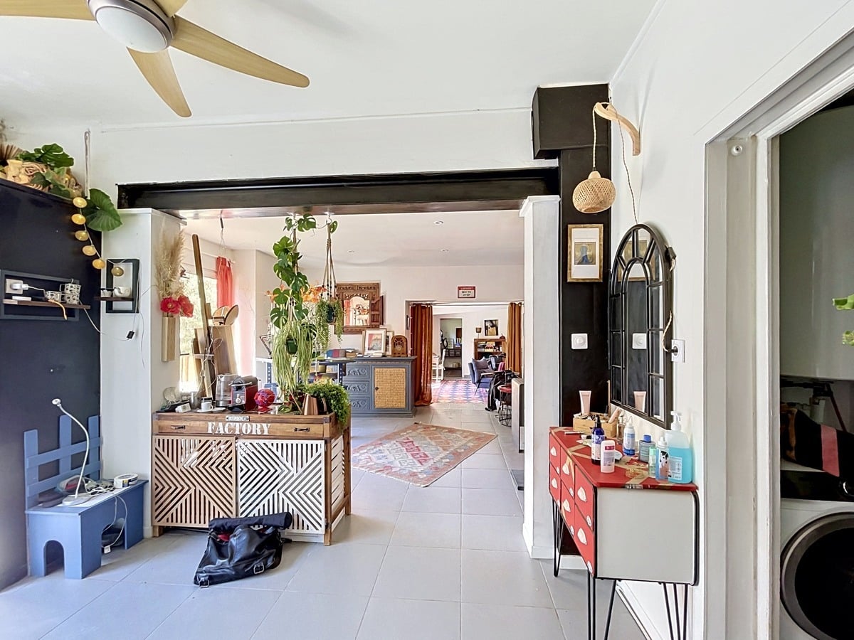 Espace de vie ouvert avec ventilateur de plafond en bois et poutre métallique noire apparente. Buffet en bois à motifs géométriques, commode rouge à tiroirs, plantes suspendues et carrelage gris clair au sol.