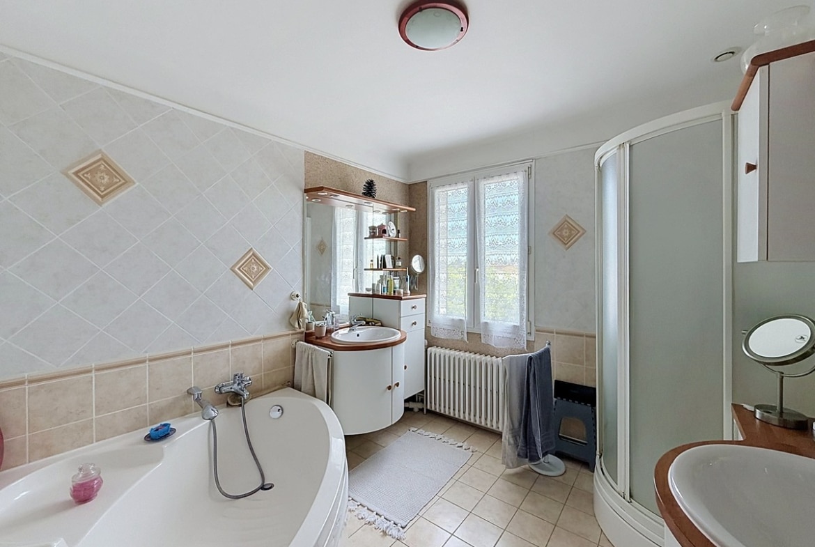 Salle de bains avec baignoire d'angle blanche et cabine de douche arrondie. Double vasque avec meuble blanc et miroir, carrelage mural beige posé en diagonale, radiateur en fonte sous la fenêtre.