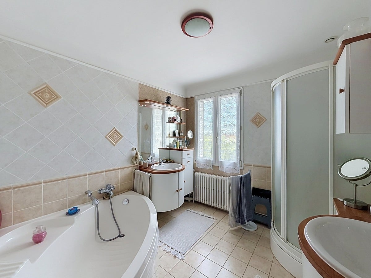 Salle de bains avec baignoire d'angle blanche et cabine de douche arrondie. Double vasque avec meuble blanc et miroir, carrelage mural beige posé en diagonale, radiateur en fonte sous la fenêtre.