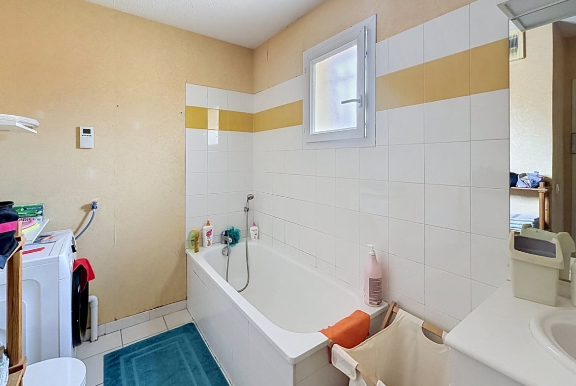 Salle de bains avec baignoire blanche encastrée contre un mur carrelé blanc et jaune. Fenêtre oscillo-battante, lavabo avec miroir, espace buanderie avec lave-linge, murs enduits jaune pâle et sol carrelé beige.