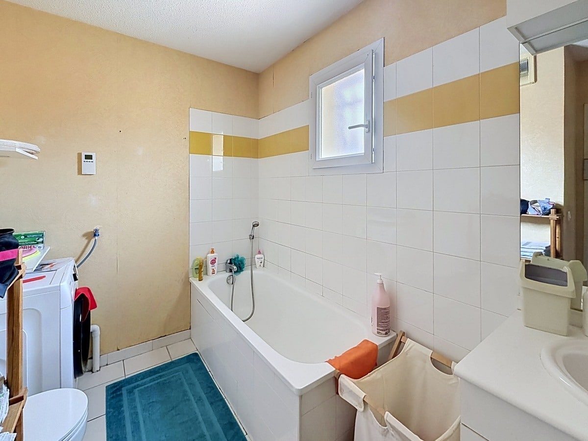 Salle de bains avec baignoire blanche encastrée contre un mur carrelé blanc et jaune. Fenêtre oscillo-battante, lavabo avec miroir, espace buanderie avec lave-linge, murs enduits jaune pâle et sol carrelé beige.