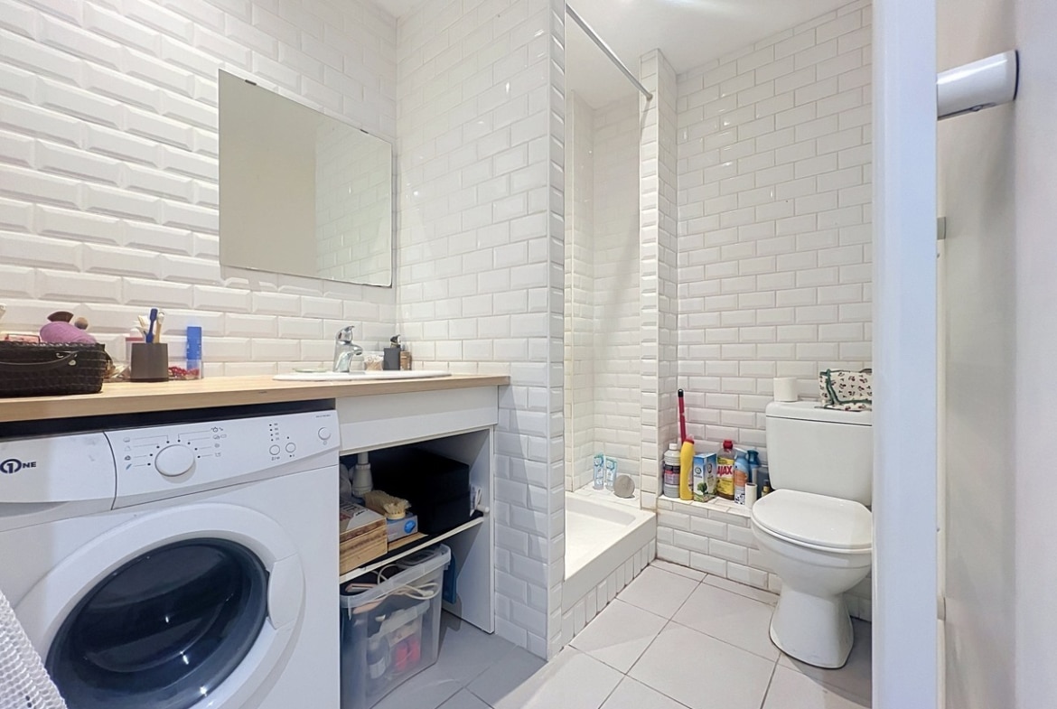 Salle de bains carrelée en faïence blanche style métro avec baignoire, WC et lave-linge intégré sous plan vasque en bois. Sol en carrelage beige clair et rangements ouverts sous le meuble vasque.