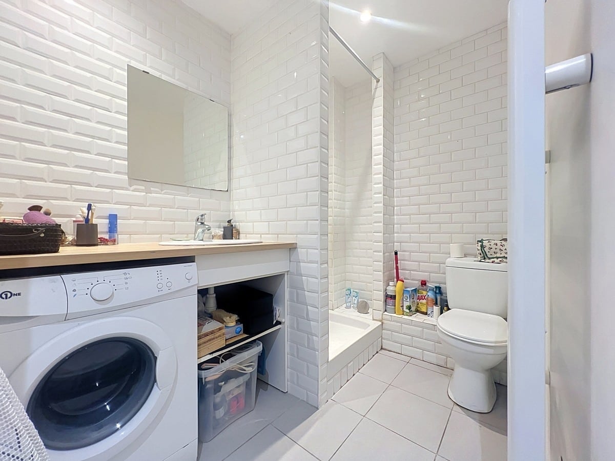 Salle de bains carrelée en faïence blanche style métro avec baignoire, WC et lave-linge intégré sous plan vasque en bois. Sol en carrelage beige clair et rangements ouverts sous le meuble vasque.