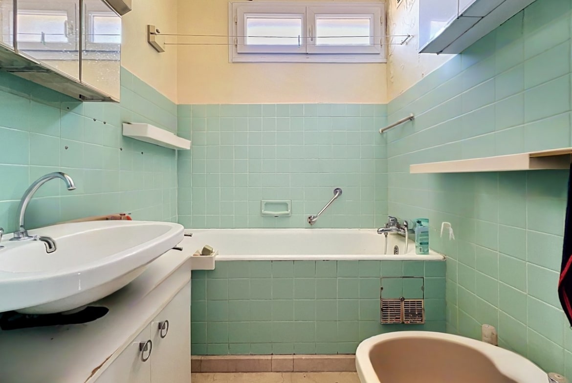 Salle de bains avec carrelage mural vert d'eau et baignoire encastrée assortie. Meuble vasque blanc avec lavabo ovale, bidet visible à droite et petite fenêtre à jalousies blanches en partie haute. Sol en vinyle beige usé.