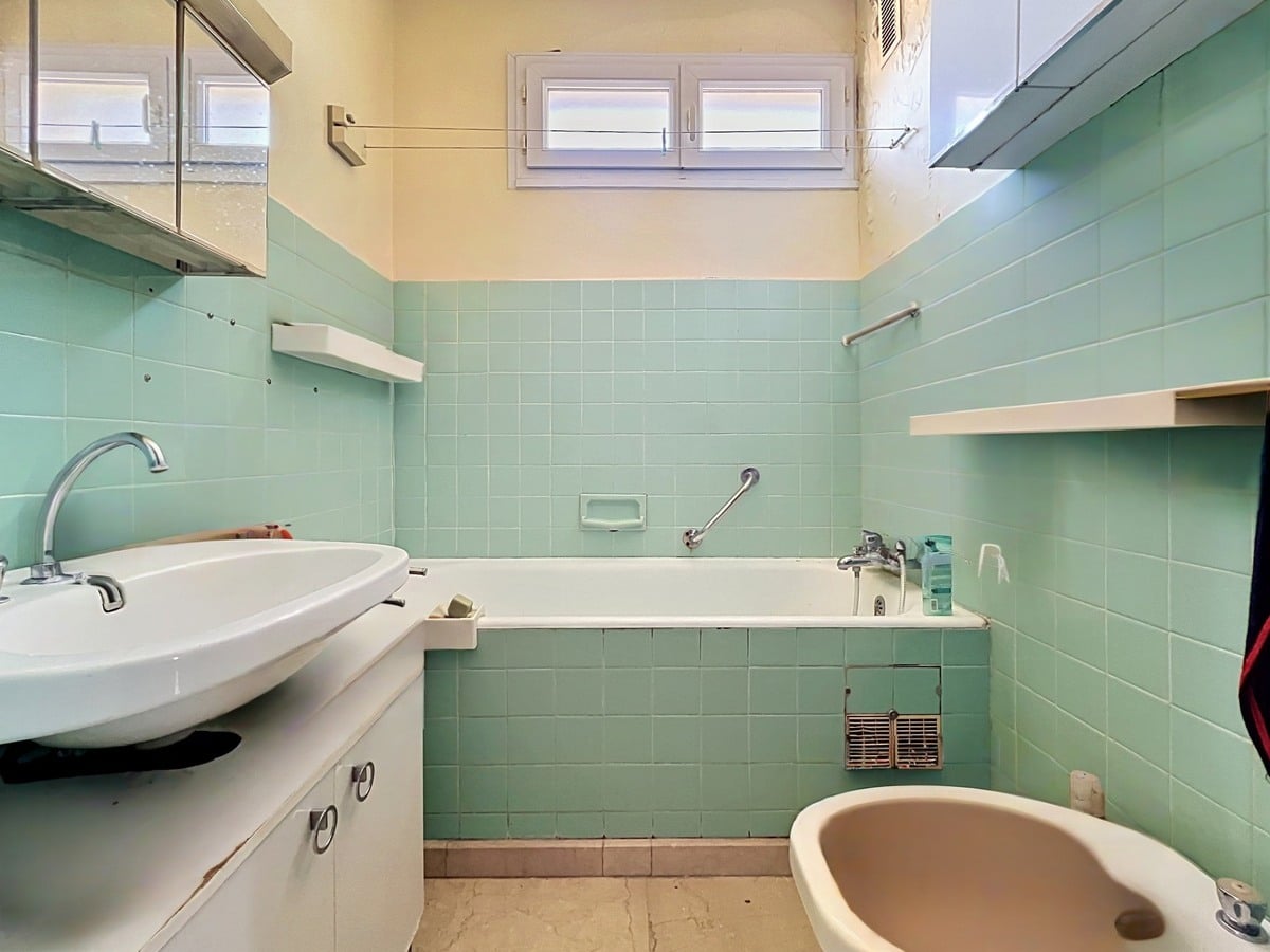 Salle de bains avec carrelage mural vert d'eau et baignoire encastrée assortie. Meuble vasque blanc avec lavabo ovale, bidet visible à droite et petite fenêtre à jalousies blanches en partie haute. Sol en vinyle beige usé.