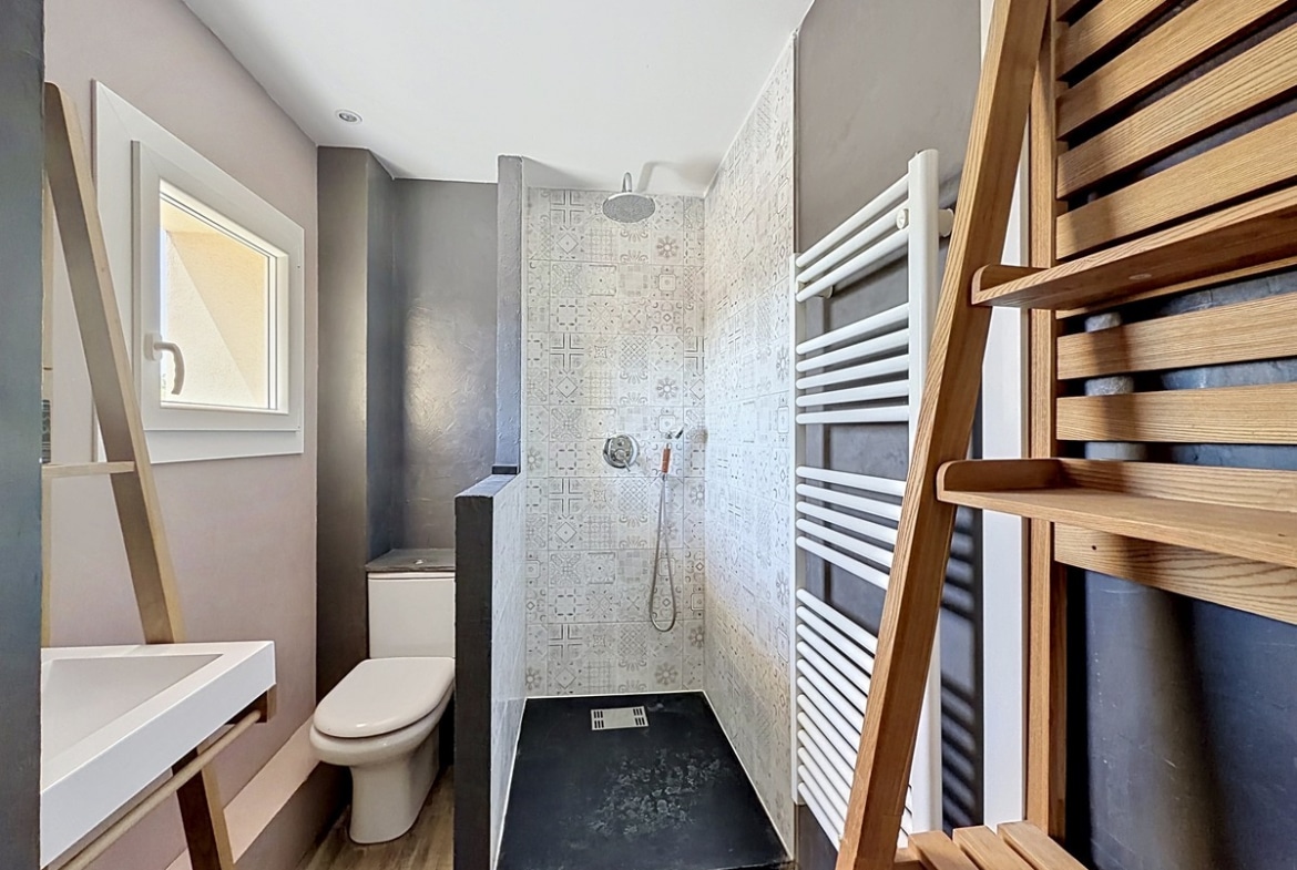 Salle d'eau avec murs gris taupe et douche à l'italienne carrelée de faïence à motifs géométriques. Vasque blanche suspendue, sèche-serviettes blanc, échelle porte-serviettes en bois.