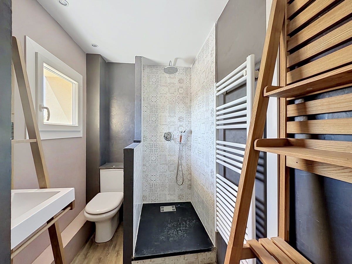 Salle d'eau avec murs gris taupe et douche à l'italienne carrelée de faïence à motifs géométriques. Vasque blanche suspendue, sèche-serviettes blanc, échelle porte-serviettes en bois.