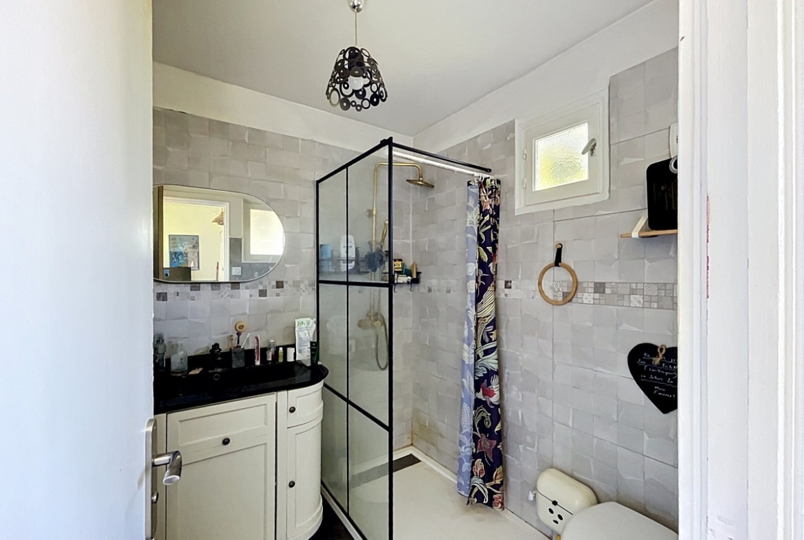 Salle de bains carrelée en gris clair avec douche à paroi vitrée noire et robinetterie dorée. Meuble vasque blanc arrondi avec plan noir, miroir ovale et WC beige visible au premier plan.