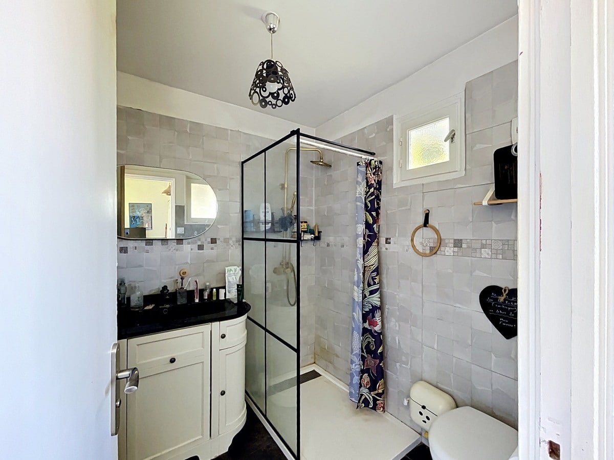 Salle de bains carrelée en gris clair avec douche à paroi vitrée noire et robinetterie dorée. Meuble vasque blanc arrondi avec plan noir, miroir ovale et WC beige visible au premier plan.
