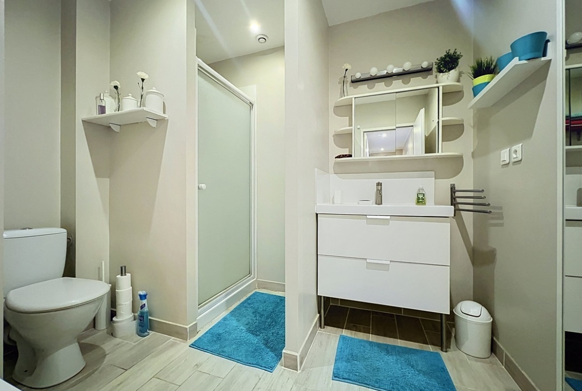 Salle de bains aux tons beige et blanc avec douche à porte vitrée opaque et WC séparé par une cloison. Meuble vasque blanc suspendu avec tiroirs, miroir avec éclairage, étagères murales et tapis bleu turquoise au sol.