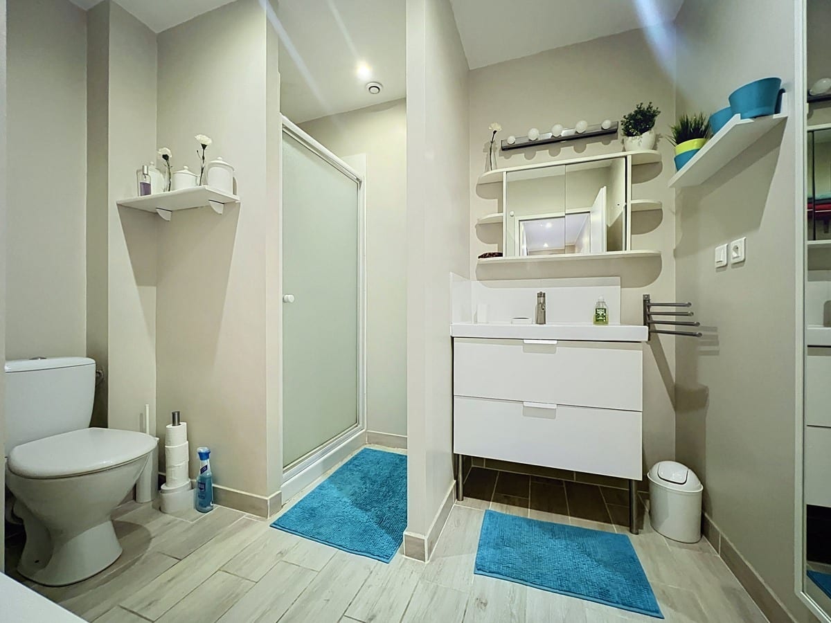Salle de bains aux tons beige et blanc avec douche à porte vitrée opaque et WC séparé par une cloison. Meuble vasque blanc suspendu avec tiroirs, miroir avec éclairage, étagères murales et tapis bleu turquoise au sol.