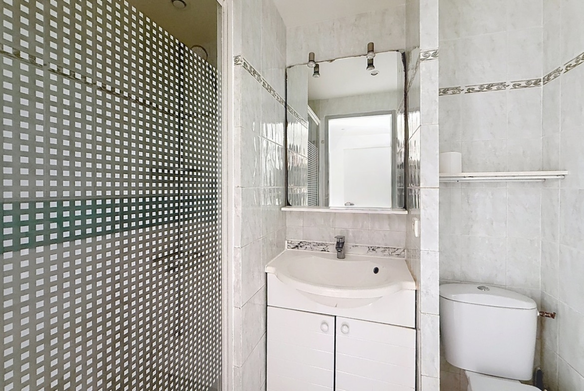 Salle d'eau carrelée en gris clair avec cabine de douche à paroi vitrée à motifs carrés. Un meuble vasque blanc surmonté d'un miroir et un WC blanc sont installés côte à côte.