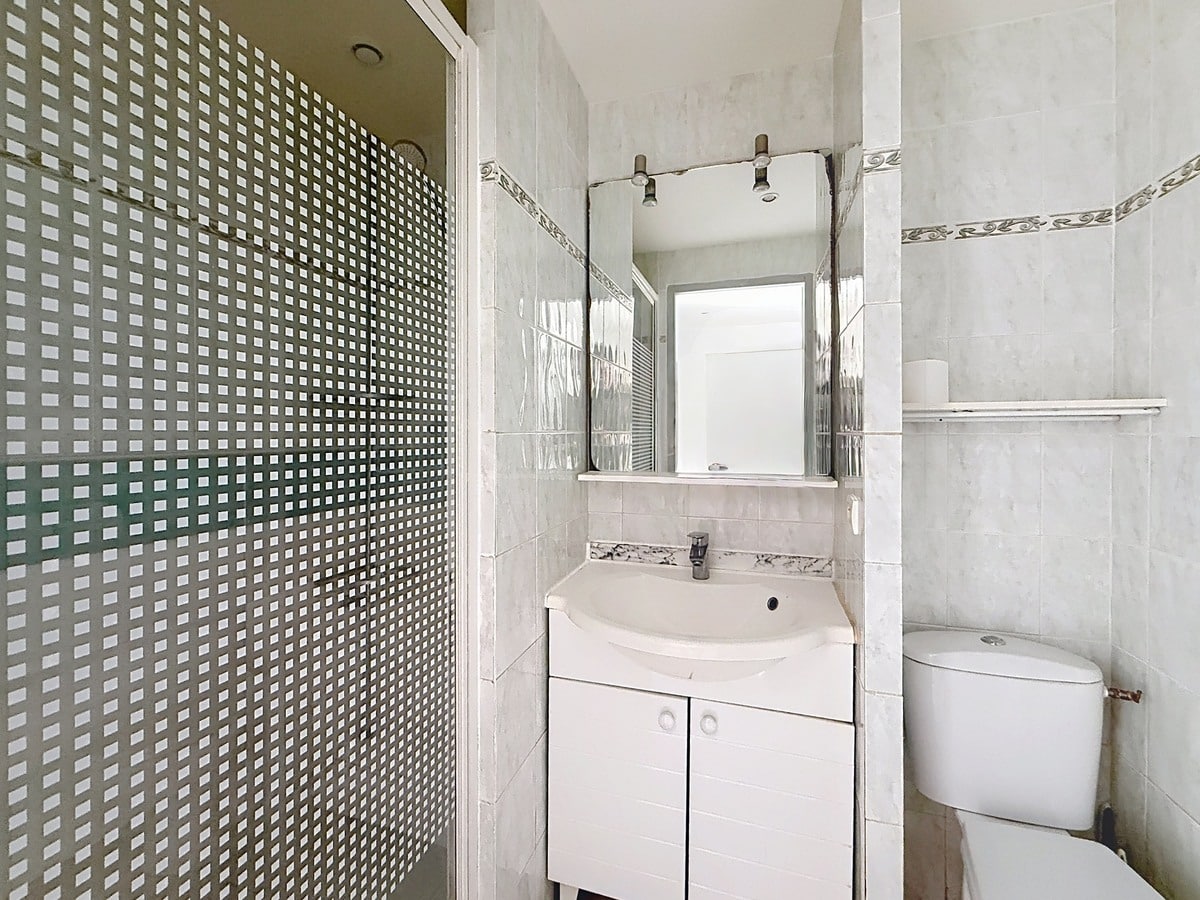 Salle d'eau carrelée en gris clair avec cabine de douche à paroi vitrée à motifs carrés. Un meuble vasque blanc surmonté d'un miroir et un WC blanc sont installés côte à côte.