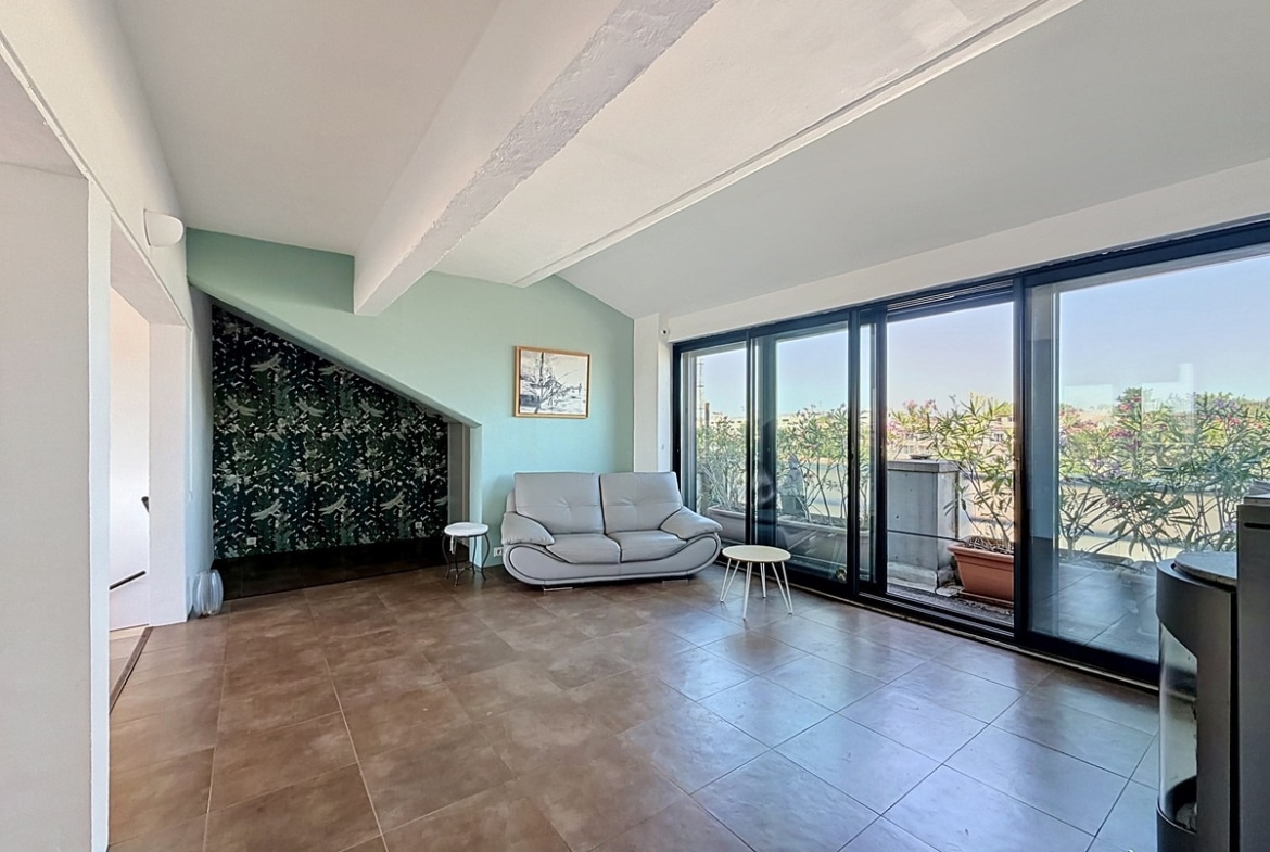 Salon avec murs vert d'eau et papier peint à motif floral sombre. Canapé gris clair, poêle à bois métallique, grandes baies vitrées noires ouvrant sur terrasse avec vue dégagée.