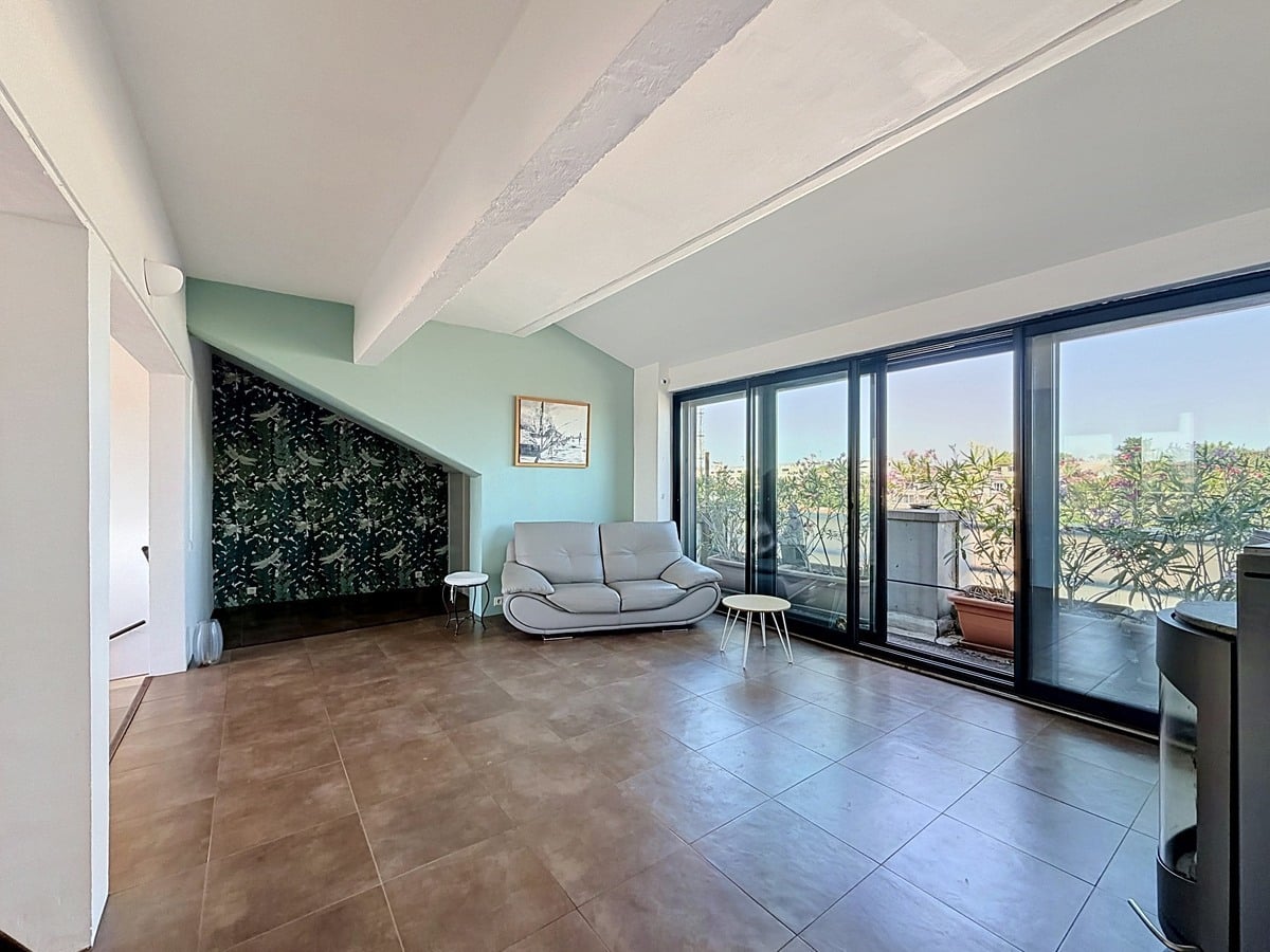 Salon avec murs vert d'eau et papier peint à motif floral sombre. Canapé gris clair, poêle à bois métallique, grandes baies vitrées noires ouvrant sur terrasse avec vue dégagée.