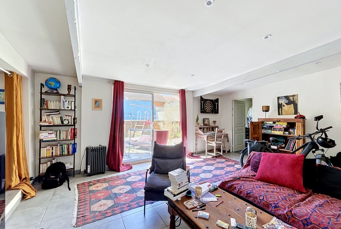Salon avec canapé rouge bordeaux et tapis à motifs géométriques rouges. Baie vitrée coulissante avec rideaux bordeaux ouvrant sur le jardin, bibliothèque en bois foncé, table basse en bois et radiateur fonte noir.