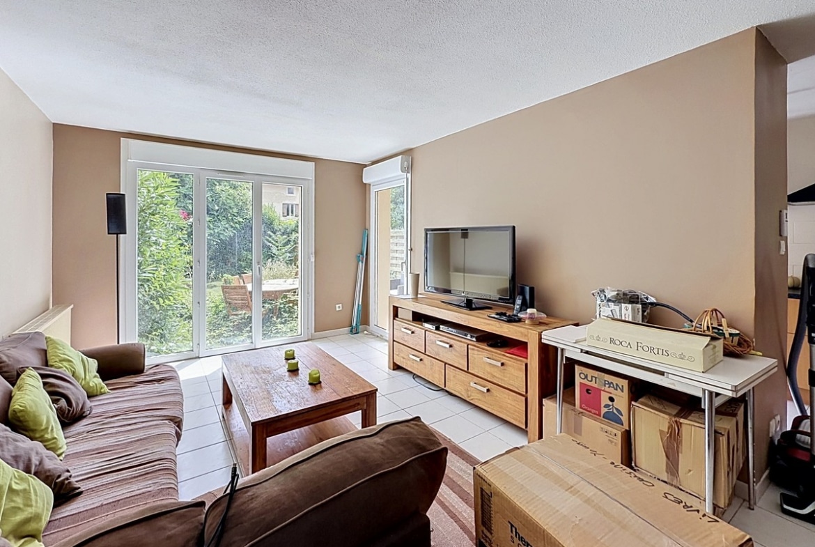 Salon avec murs beige et sol carrelé blanc, meublé d'un canapé marron et d'un meuble TV en bois massif avec tiroirs. Grande baie vitrée à double vantaux donnant sur le jardin verdoyant, cartons de déménagement