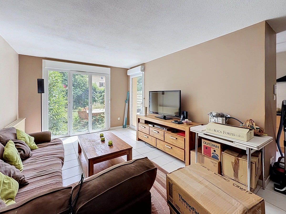 Salon avec murs beige et sol carrelé blanc, meublé d'un canapé marron et d'un meuble TV en bois massif avec tiroirs. Grande baie vitrée à double vantaux donnant sur le jardin verdoyant, cartons de déménagement