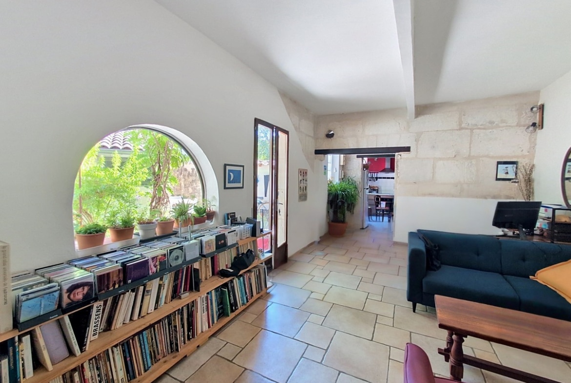 Séjour avec étagères en bois remplies de livres et vinyles, surmonté d'une fenêtre en demi-cercle avec plantes vertes. Canapé bleu canard avec coussin jaune, sol en carrelage beige et murs en pierre apparente blanche.