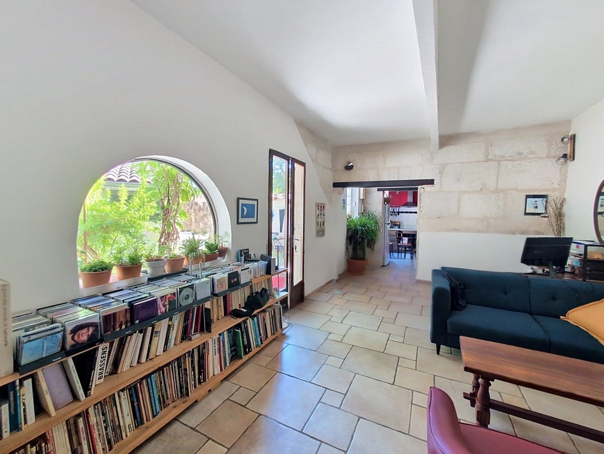 Séjour avec étagères en bois remplies de livres et vinyles, surmonté d'une fenêtre en demi-cercle avec plantes vertes. Canapé bleu canard avec coussin jaune, sol en carrelage beige et murs en pierre apparente blanche.