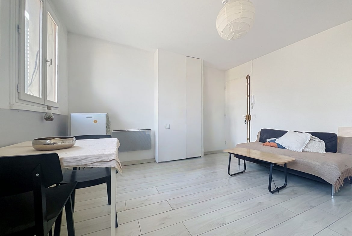 Pièce de vie avec canapé convertible beige et bleu marine, table basse en bois et métal noir. Radiateur électrique mural blanc, placard intégré et sol en parquet stratifié gris clair.