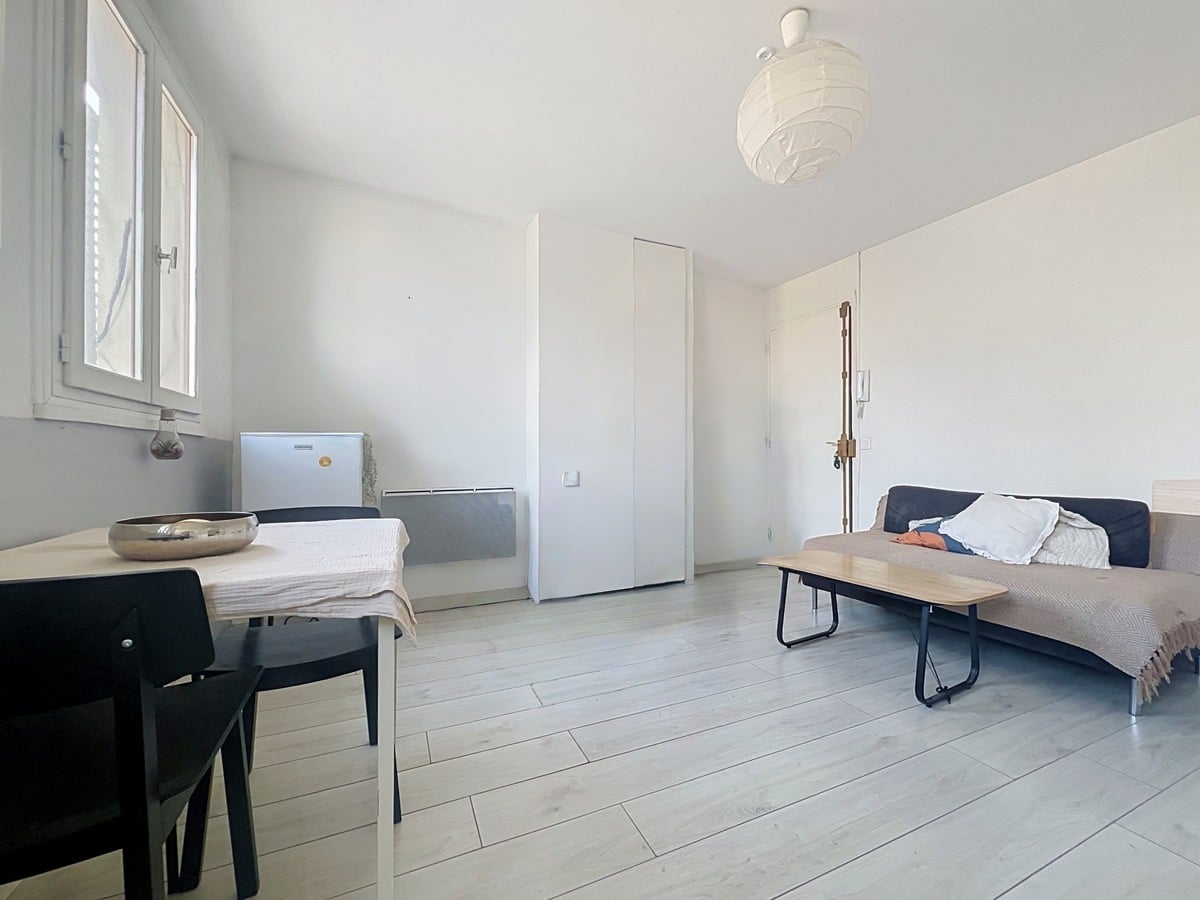 Pièce de vie avec canapé convertible beige et bleu marine, table basse en bois et métal noir. Radiateur électrique mural blanc, placard intégré et sol en parquet stratifié gris clair.