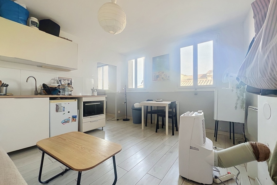 Pièce de vie avec kitchenette blanche équipée de réfrigérateur et four, sol en parquet stratifié clair. Trois fenêtres à petits carreaux côté sud, table noire avec chaises et climatiseur mobile visible.