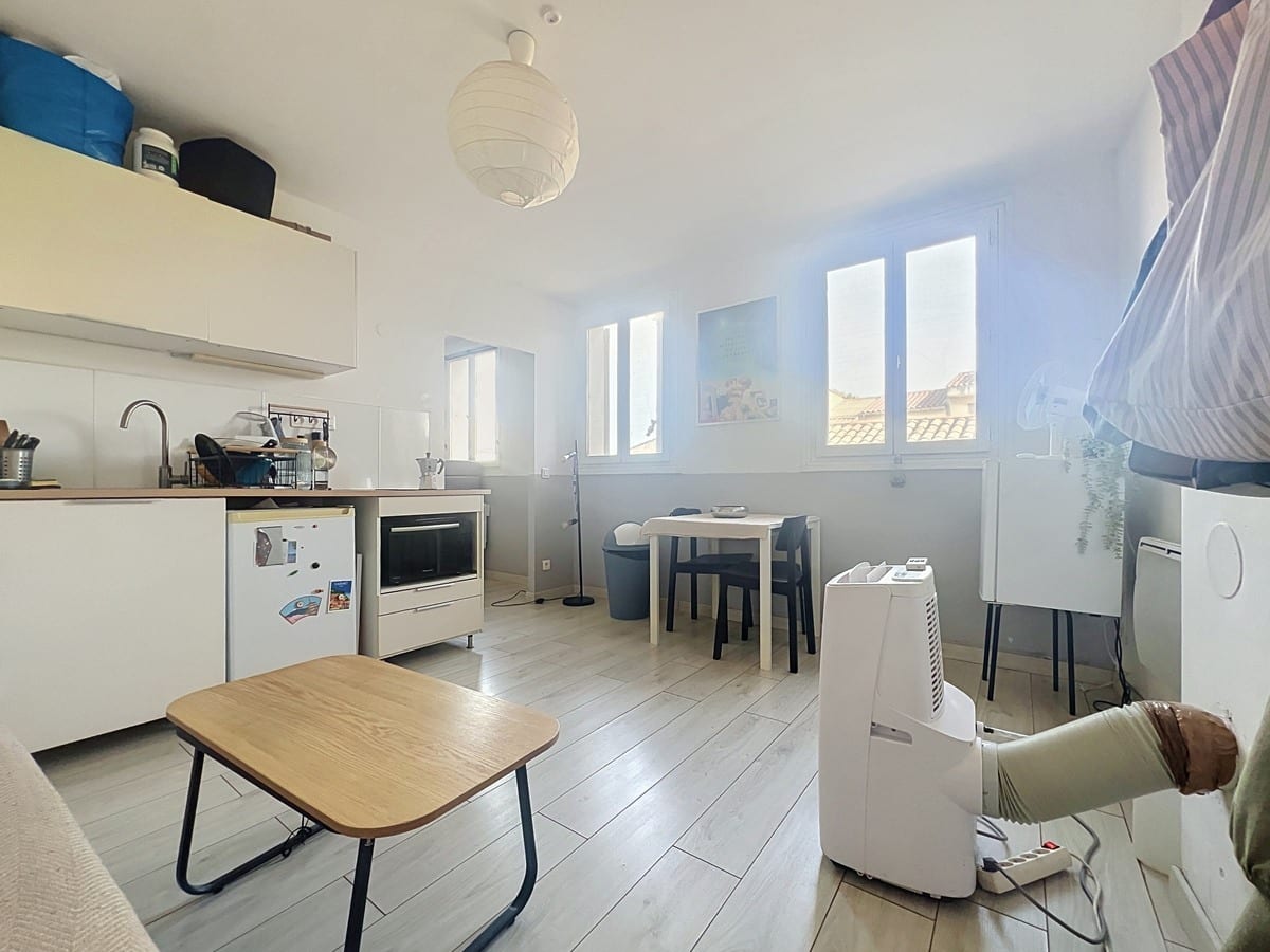 Pièce de vie avec kitchenette blanche équipée de réfrigérateur et four, sol en parquet stratifié clair. Trois fenêtres à petits carreaux côté sud, table noire avec chaises et climatiseur mobile visible.