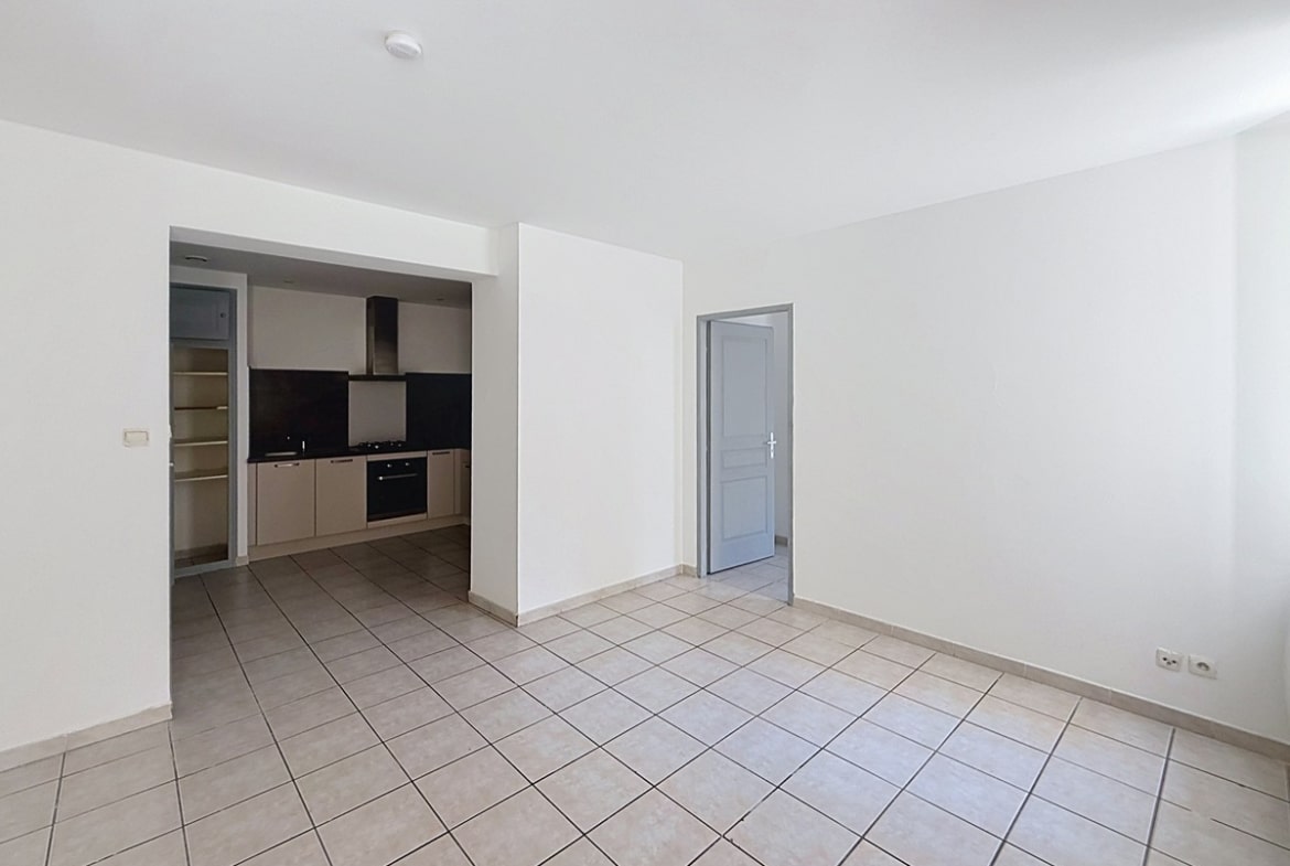 Séjour vide avec carrelage beige et murs blancs donnant sur une cuisine équipée en alcôve. La cuisine présente des meubles bas taupe, un plan de travail noir, une hotte aspirante et un four encastré. Photo 2 — salle-de-bain-appartement-avignon-vente-p2-web.jpg