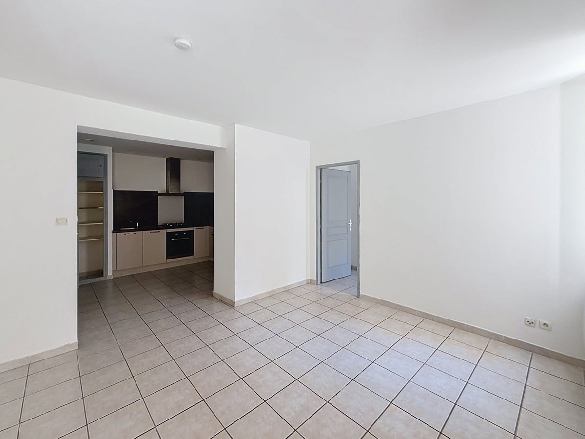 Séjour vide avec carrelage beige et murs blancs donnant sur une cuisine équipée en alcôve. La cuisine présente des meubles bas taupe, un plan de travail noir, une hotte aspirante et un four encastré. Photo 2 — salle-de-bain-appartement-avignon-vente-p2-web.jpg
