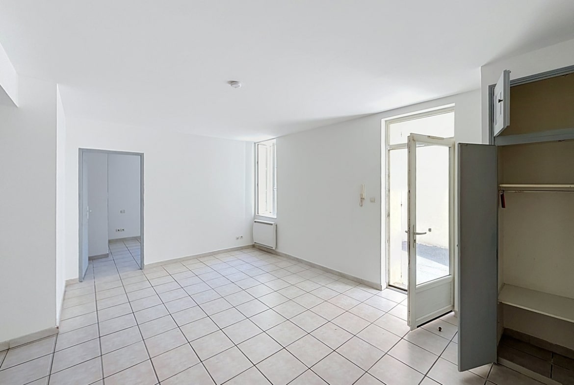 Pièce de vie vide avec sol carrelé beige et murs blancs, porte d'entrée vitrée ouverte sur la rue. Un placard avec étagères intégrées est visible à droite de l'entrée.