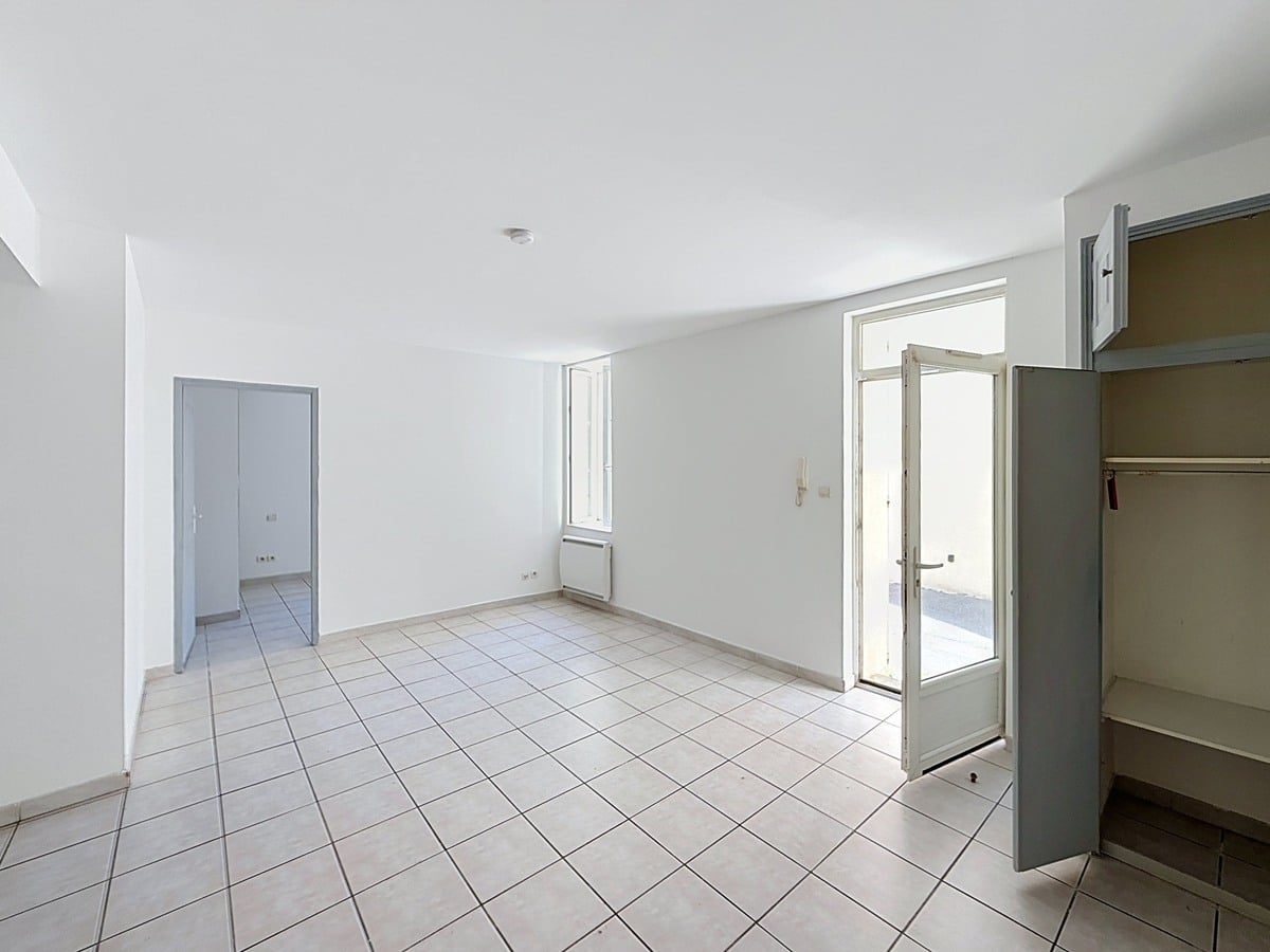 Pièce de vie vide avec sol carrelé beige et murs blancs, porte d'entrée vitrée ouverte sur la rue. Un placard avec étagères intégrées est visible à droite de l'entrée.