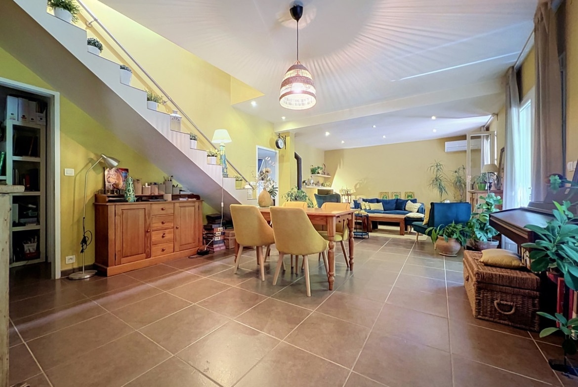Grand séjour avec murs jaunes et carrelage beige, escalier intérieur avec rampe métallique et plantes décoratives. Table en bois massif avec chaises jaunes, buffet en pin, canapé bleu en arrière-plan et climatisation murale visible.