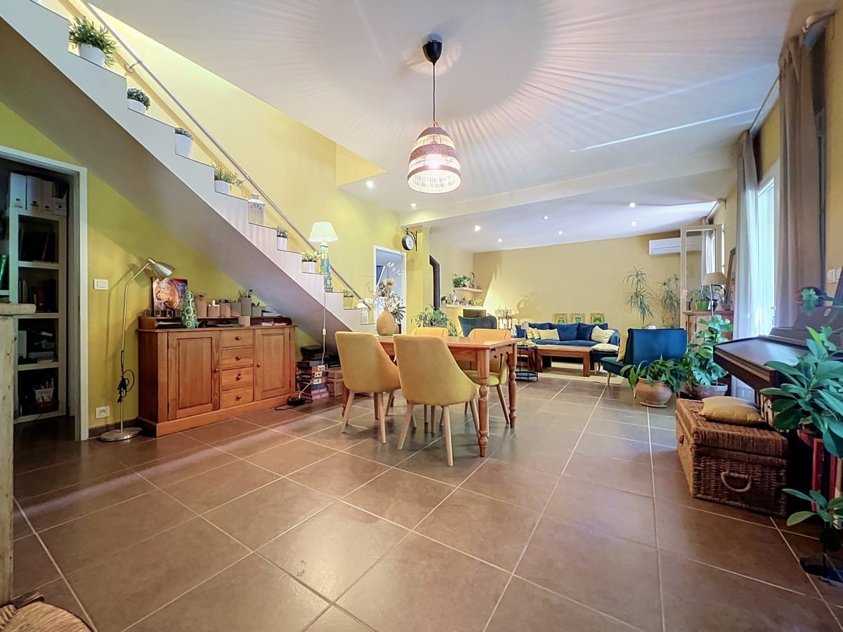Grand séjour avec murs jaunes et carrelage beige, escalier intérieur avec rampe métallique et plantes décoratives. Table en bois massif avec chaises jaunes, buffet en pin, canapé bleu en arrière-plan et climatisation murale visible.