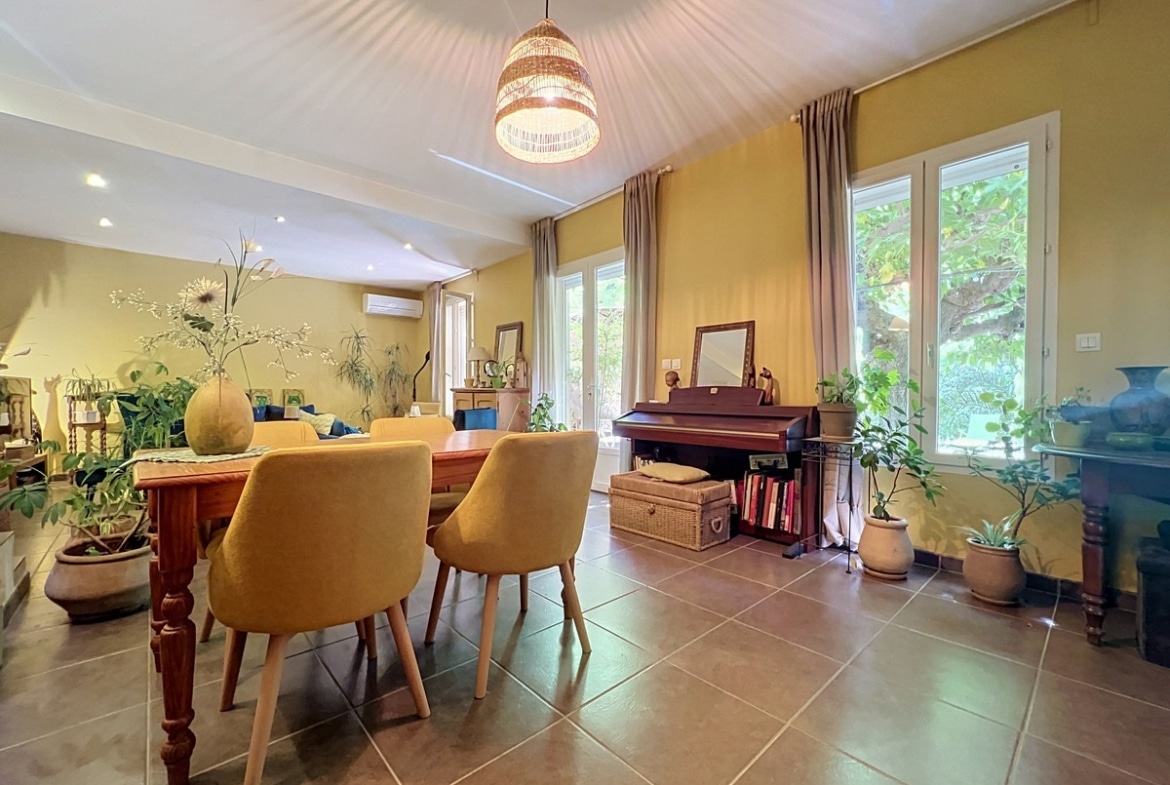 Séjour lumineux avec hauts plafonds lambris blanc et murs jaunes, trois grandes fenêtres avec rideaux taupe. Piano droit en bois foncé, table de repas avec chaises jaunes, nombreuses plantes vertes et malle en osier.
