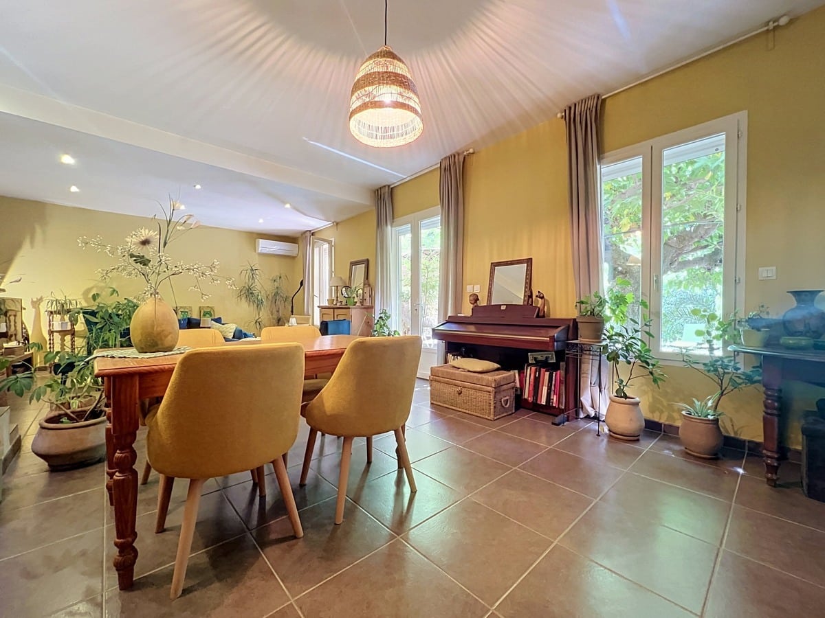Séjour lumineux avec hauts plafonds lambris blanc et murs jaunes, trois grandes fenêtres avec rideaux taupe. Piano droit en bois foncé, table de repas avec chaises jaunes, nombreuses plantes vertes et malle en osier.