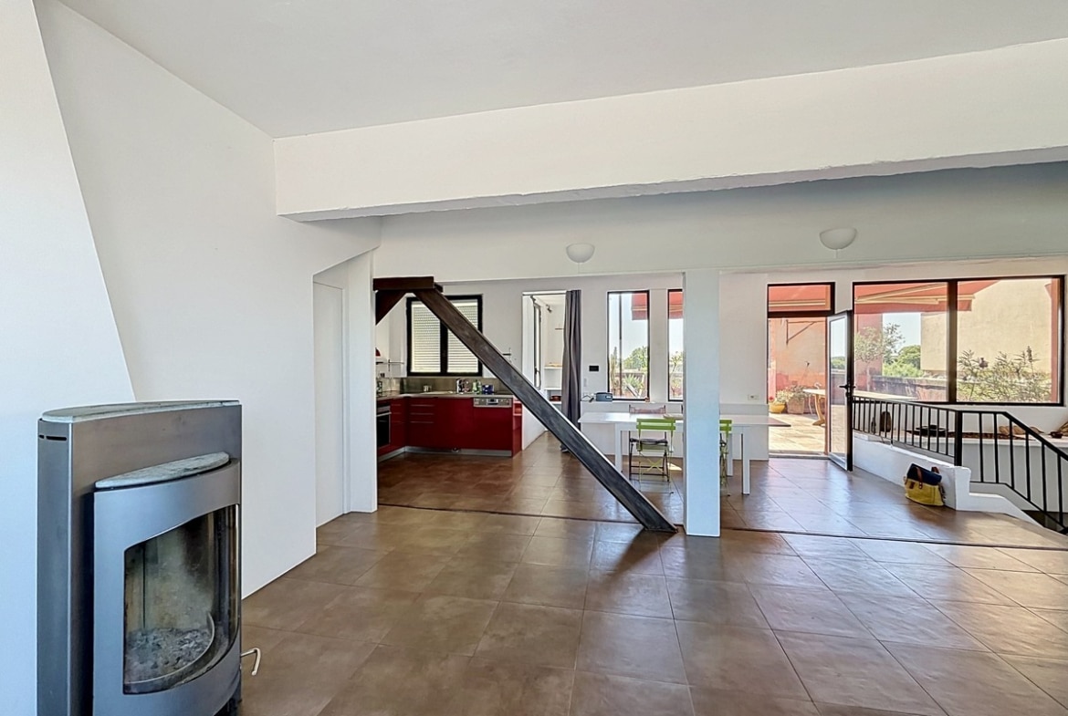 Vaste séjour en duplex avec carrelage marron et poutre décorative en bois. Poêle à bois gris métallique, escalier avec garde-corps en fer forgé, cuisine ouverte visible en arrière-plan.