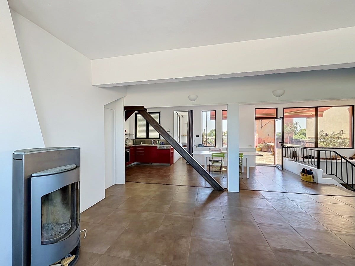 Vaste séjour en duplex avec carrelage marron et poutre décorative en bois. Poêle à bois gris métallique, escalier avec garde-corps en fer forgé, cuisine ouverte visible en arrière-plan.