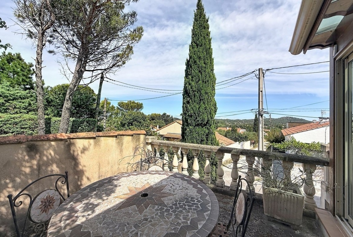 Terrasse extérieure avec table ronde en mosaïque grise et chaises en fer forgé noir. Vue dégagée sur les toits en tuiles et cyprès, balustrade à balustres blancs et mur en crépi beige.